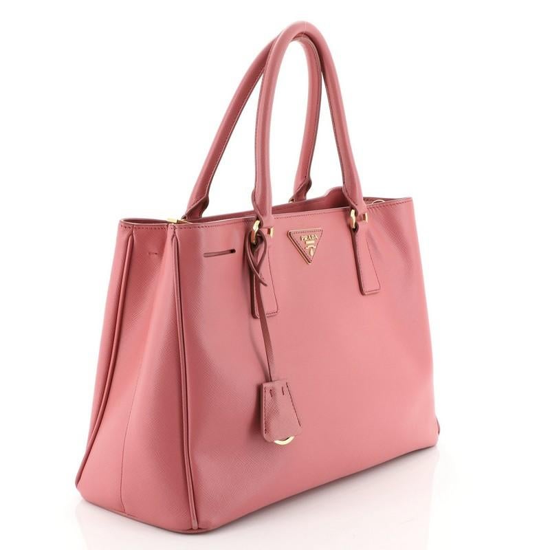 This Prada Gardener's Tote Saffiano Leather Medium, crafted from pink saffiano leather, features dual rolled leather handles, raised Prada logo plate, and gold-tone hardware. Its magnetic snap closure opens to a pink fabric interior with middle zip