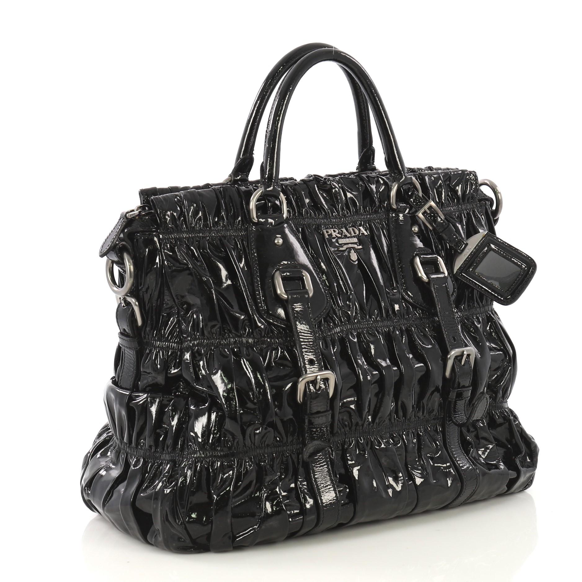 This Prada Gaufre Convertible Tote Patent Medium, crafted in black patent leather, features dual rolled handles with belt and buckle detailing and gunmetal-tone hardware. Its zip closure opens to a black fabric interior with side zip and slip