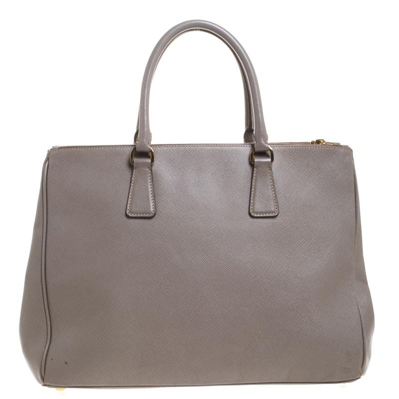 Feminine in shape and grand in design, this Double Zip tote by Prada will be a loved addition to your closet. It has been crafted from grey Saffiano Lux leather and styled minimally with gold-tone hardware. It comes with two top handles, two zip
