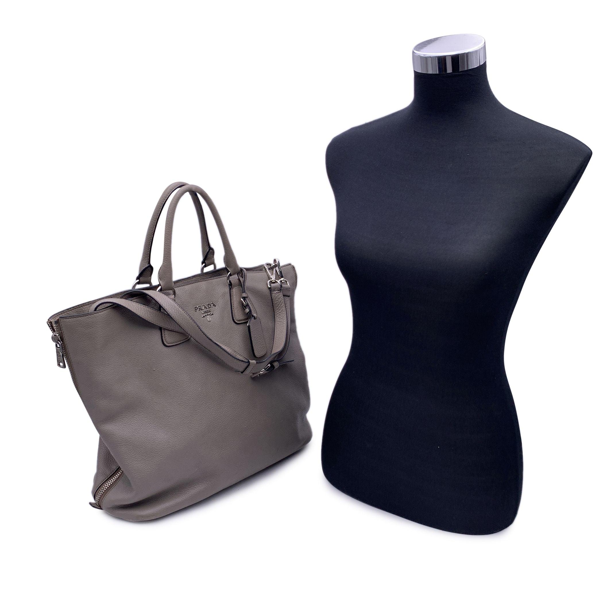 Beautiful PRADA tote bag in Grey Taupe textured leather. It features double round top handles and adjustable and removable shoulder strap. Prada lettering on the front in silver metal. Upper zipper closure. 4 bottom feet. The bag has 1 main open