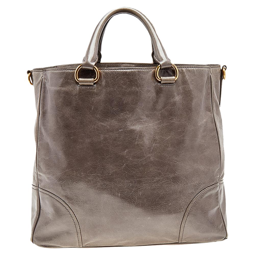 This grey bag from Prada is very chic and stylish. It is crafted from Vitello shine leather that looks amazing. It features dual top handles with an attached tag accent, a gold-tone brand logo at the front, and protective metal feet. It also comes