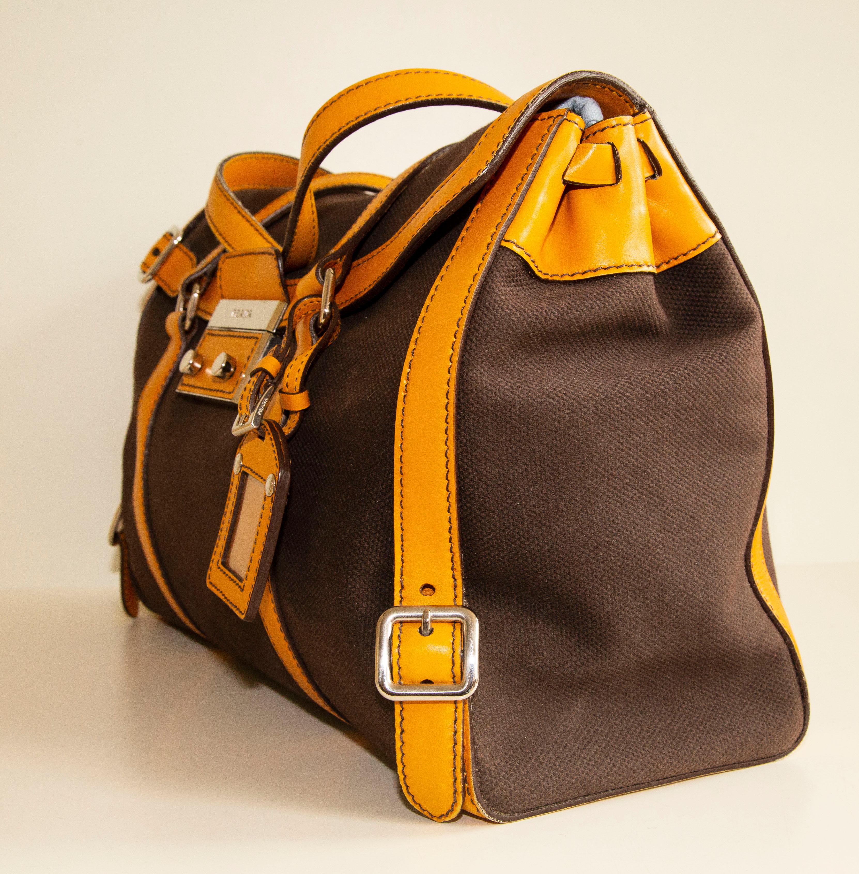 An authentic Prada shoulder bag. The bag features a brown canvas exterior, mustard yellow leather trim, and silver-tone hardware. The interior is lined with yellow leather and next to the major compartiment, there is one zipped pocket.  Tha bag is