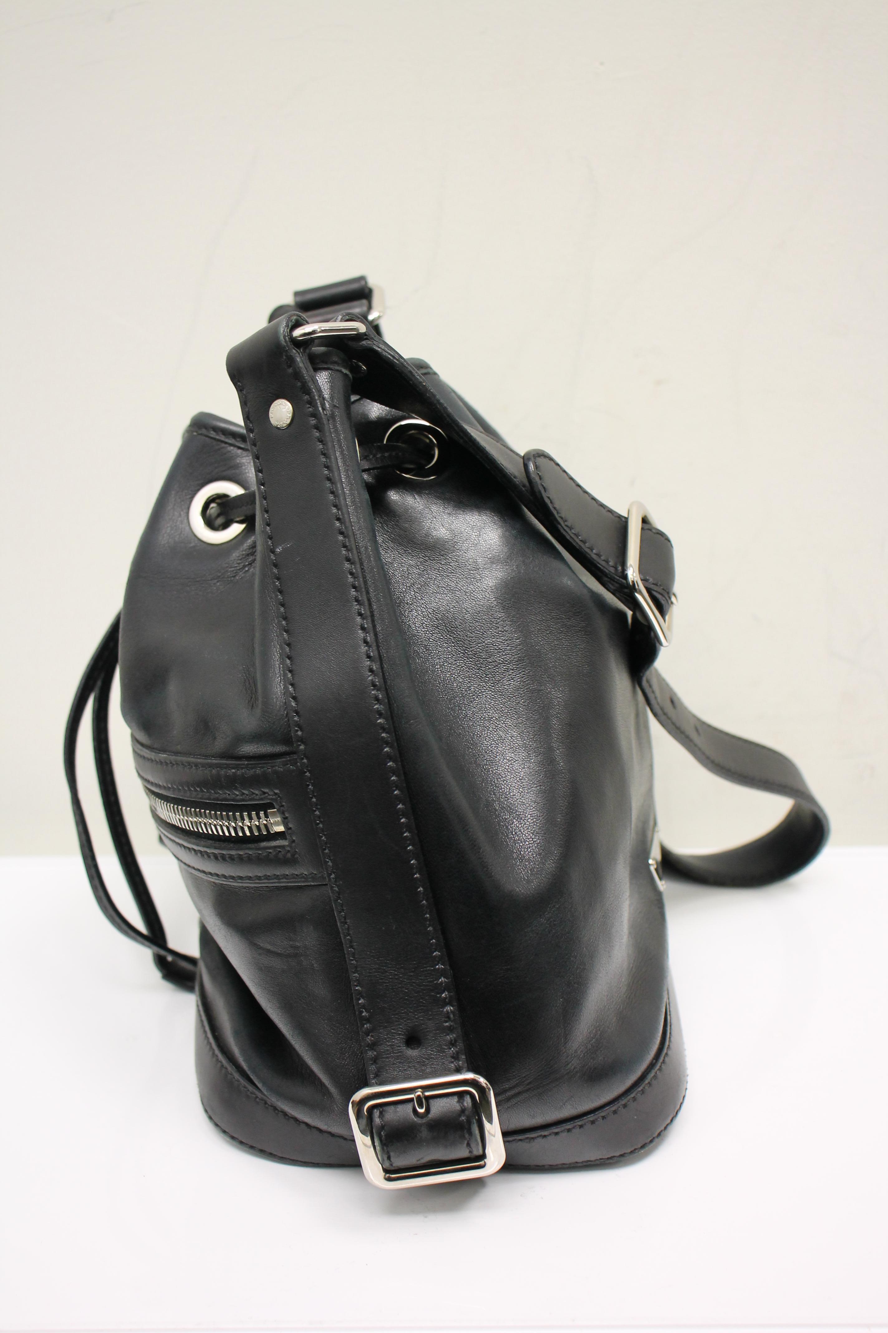Black leather. Silver-tone hardware. Pull closure at top. Adjustable flat shoulder strap. Logo at back. Buckle detail at sides. One exterior zippered pocket at front. Lined interior. One interior zippered pocket.