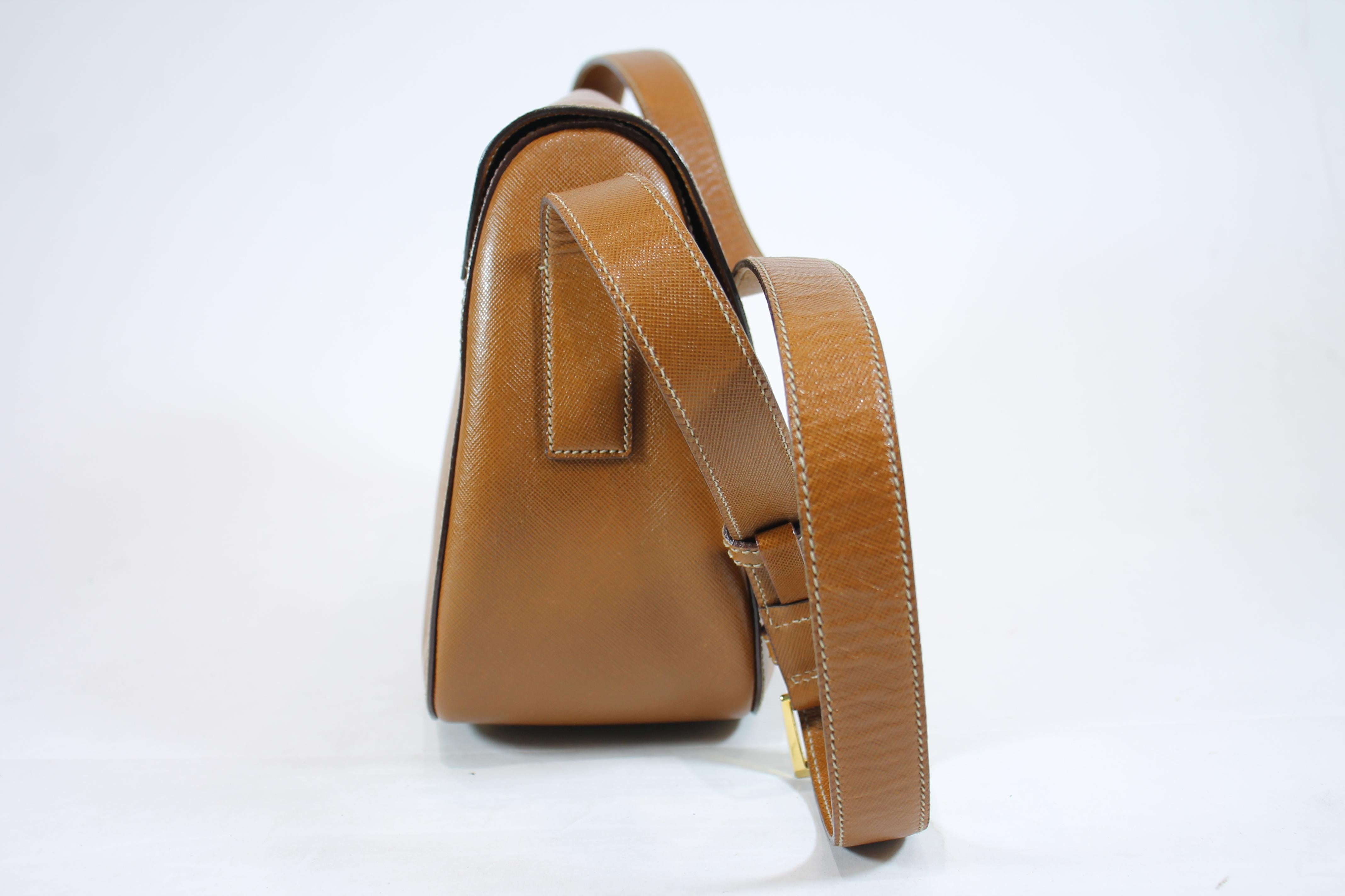 Brown camel grained leather. Gold-tone hardware. Front flap with push lock closure with prada engraved logo. Adjustable flat shoulder strap. One interior zippered pocket. Protective feet at base.