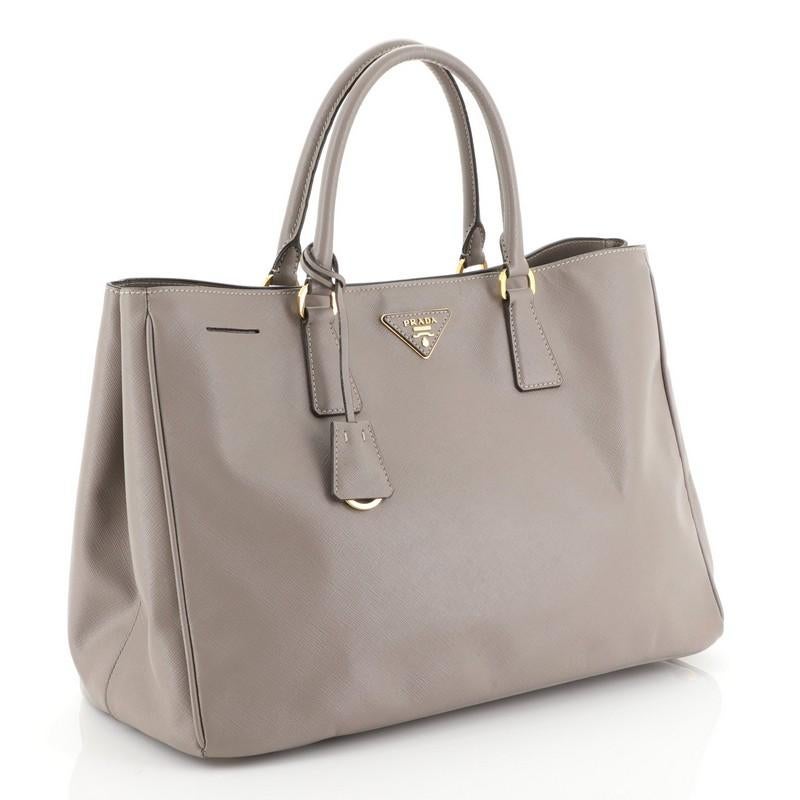 This Prada Lux Open Tote Saffiano Leather Large, crafted from neutral saffiano leather, features dual rolled handles, side snap buttons, protective base studs, and gold-tone hardware. Its magnetic snap closure opens to a brown fabric interior with