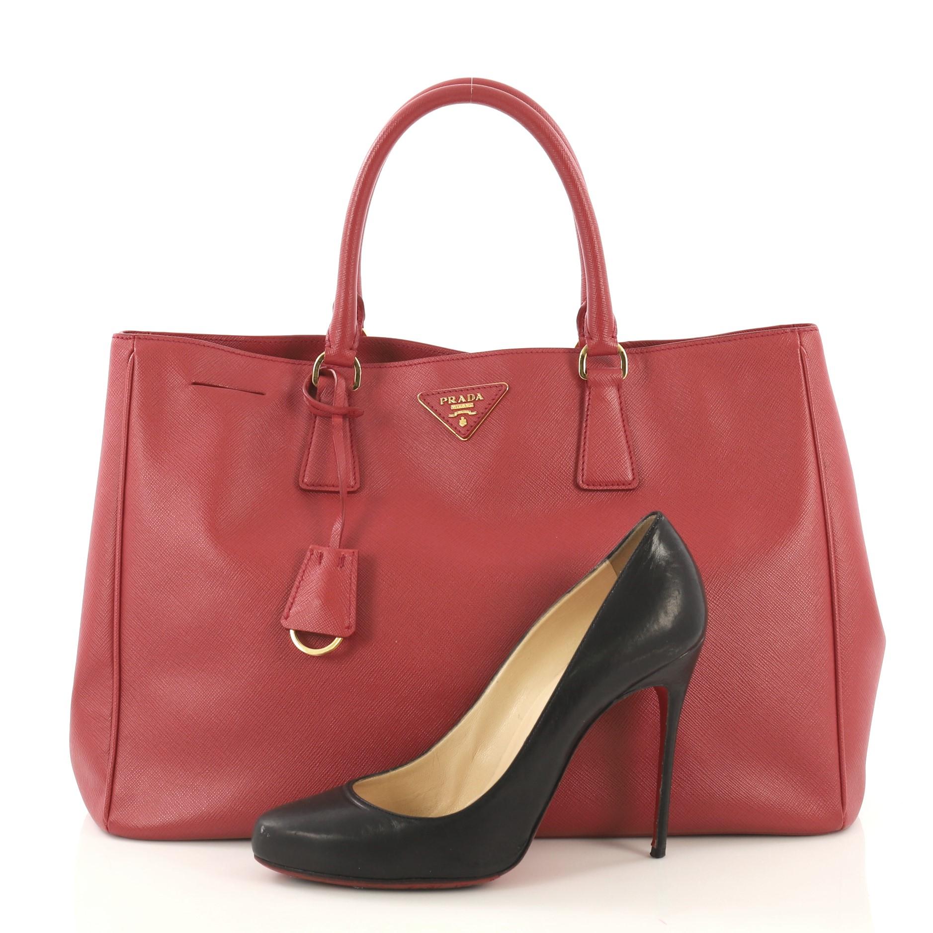 This Prada Lux Open Tote Saffiano Leather Medium, crafted from red saffiano leather, features dual rolled handles, side snap buttons, protective base studs, and gold-tone hardware. Its magnetic snap closure opens to a red fabric interior with side