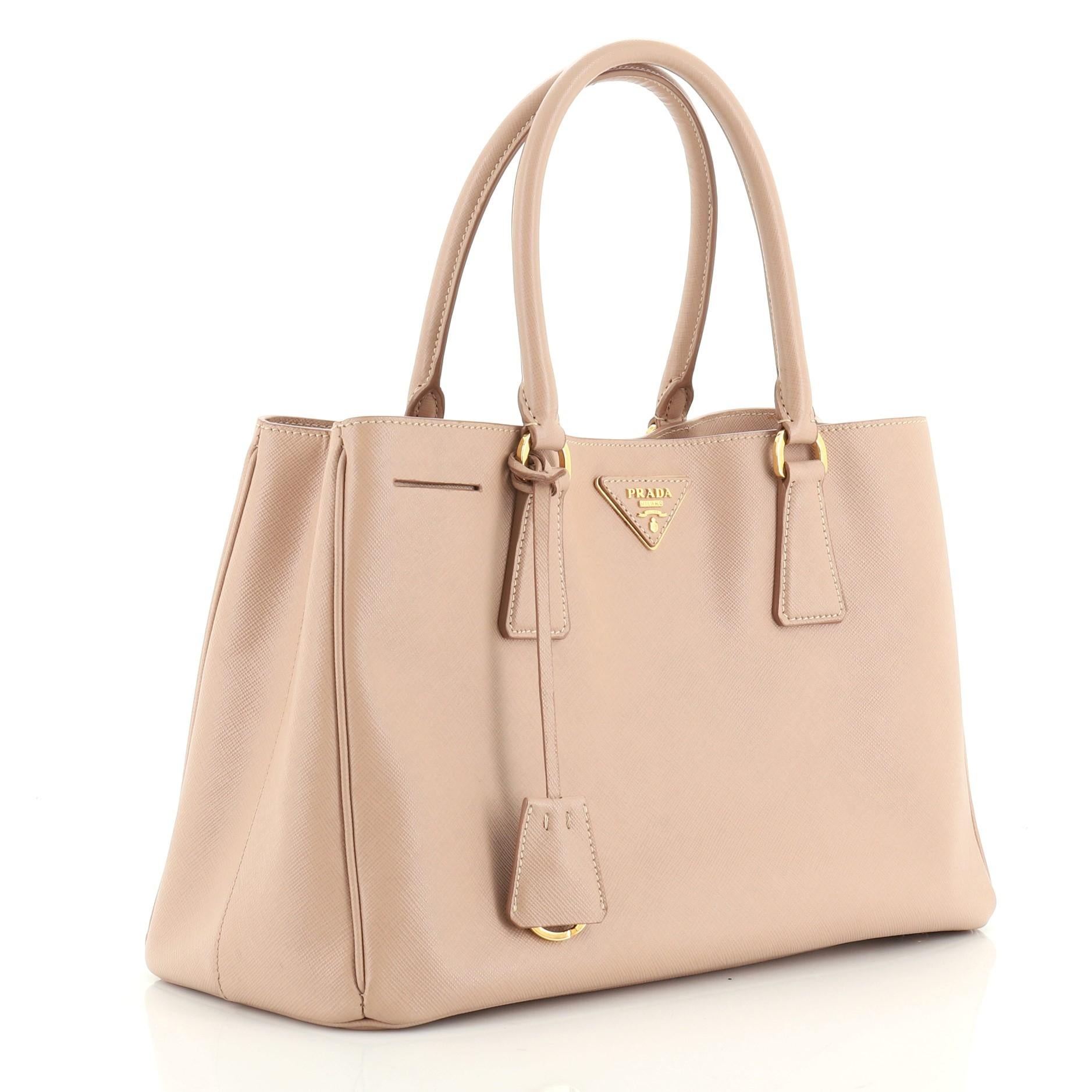 This Prada Lux Open Tote Saffiano Leather Small, crafted from neutral saffiano leather, features dual rolled handles, side snap buttons, protective base studs, and gold-tone hardware. Its magnetic snap closure opens to a brown fabric interior with