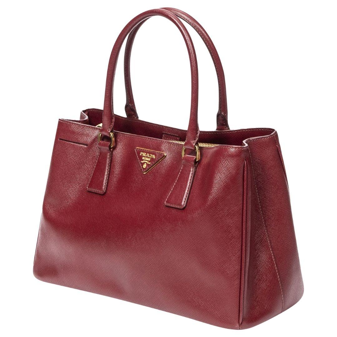 A sophisticated maroon Saffiano leather tote with gold hardware and a magnetic snap closure. It opens to a leather interior with two compartments, two zippered pockets, and one slip pocket for organized storage.

SPECIFICS
Length: 13
Width: