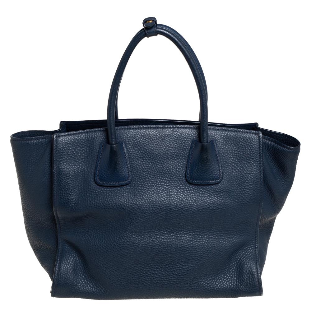 This Front Pocket Wing tote by Prada is an investment-worthy creation. It is constructed using Vitello Daino leather in navy blue and equipped with two handles, a front zip pocket, and gold-tone hardware. It has a nylon-lined interior with a zip