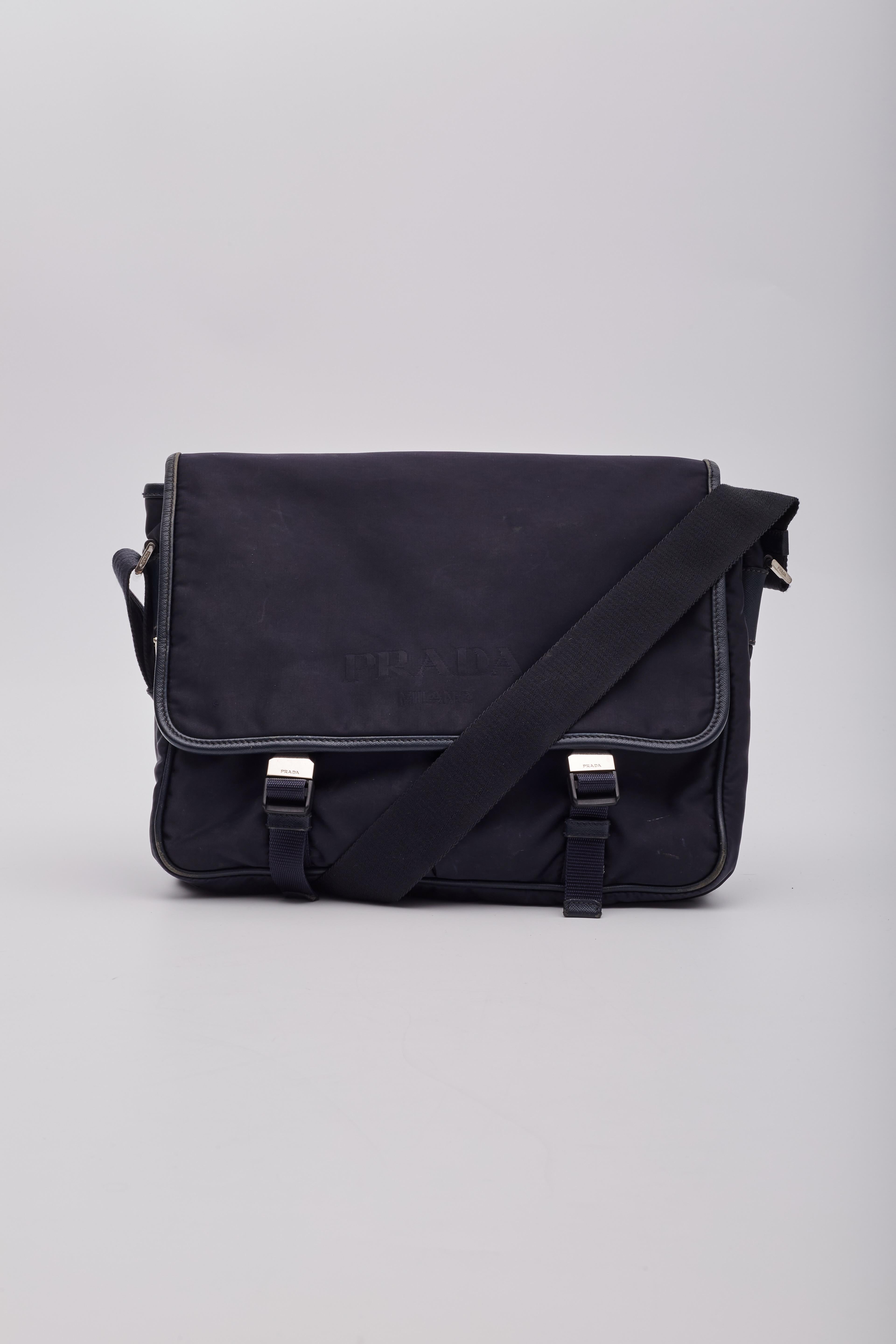 The Prada messenger bag is made of black Tessuto nylon with saffiano leather trim embellishments. The bag features a single exterior pocket, a large flap with double buckle closure at the front and silver tone hardware. The bag is finished with logo