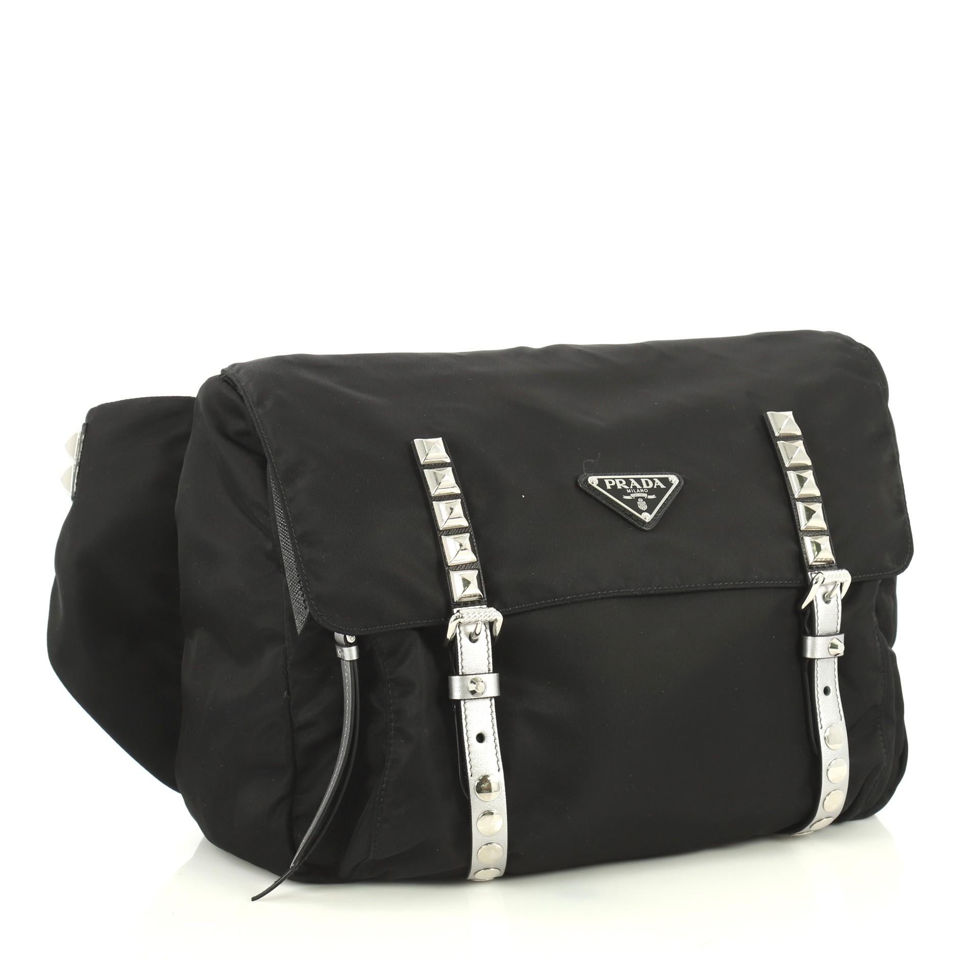 This Prada New Vela Flap Messenger Bag Tessuto with Studded Leather Medium, crafted in black tessuto with studded leather, features an adjustable leather strap, exterior back zip pocket, and silver-tone hardware. Its buckle and zip closure opens to