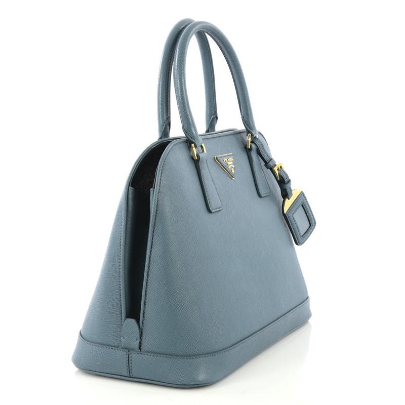 This Prada Open Promenade Bag Saffiano Leather Large, crafted in light blue saffiano leather, features dual rolled handles, protective base studs, Prada logo at the center, and gold-tone hardware. Its magnetic snap closure opens to a light blue