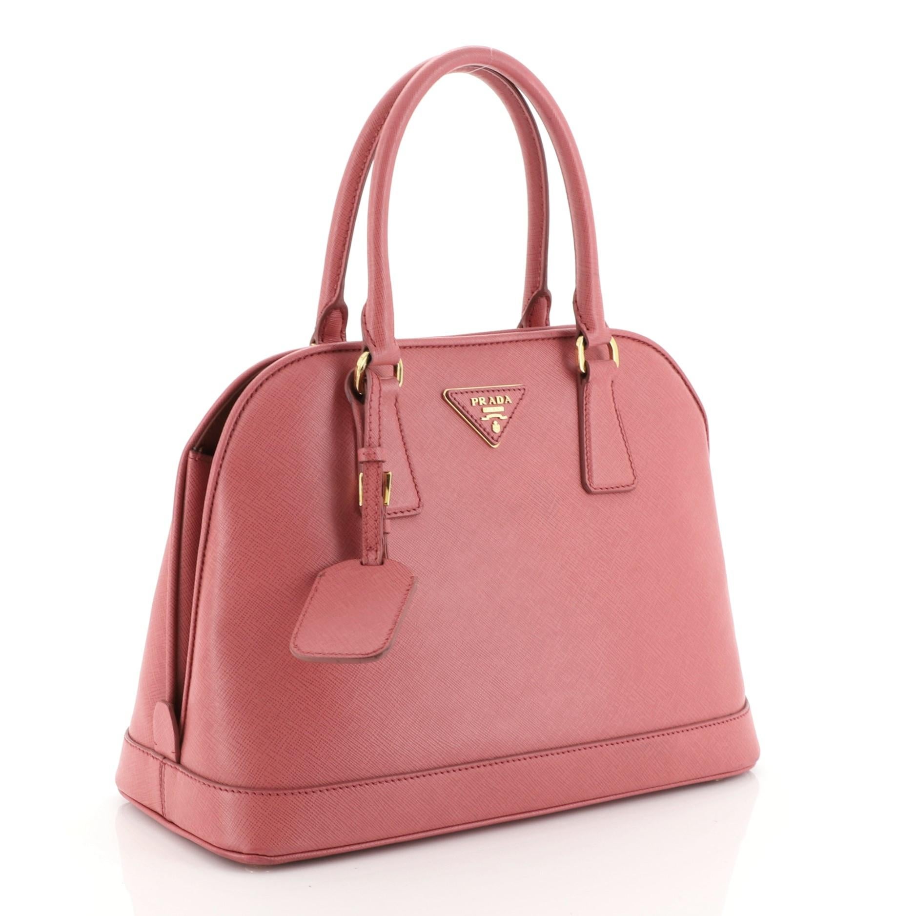 This Prada Open Promenade Bag Saffiano Leather Medium, crafted in pink saffiano leather, features dual rolled handles, protective base studs, Prada logo at the center, and gold-tone hardware. Its magnetic snap button closure opens to a pink fabric