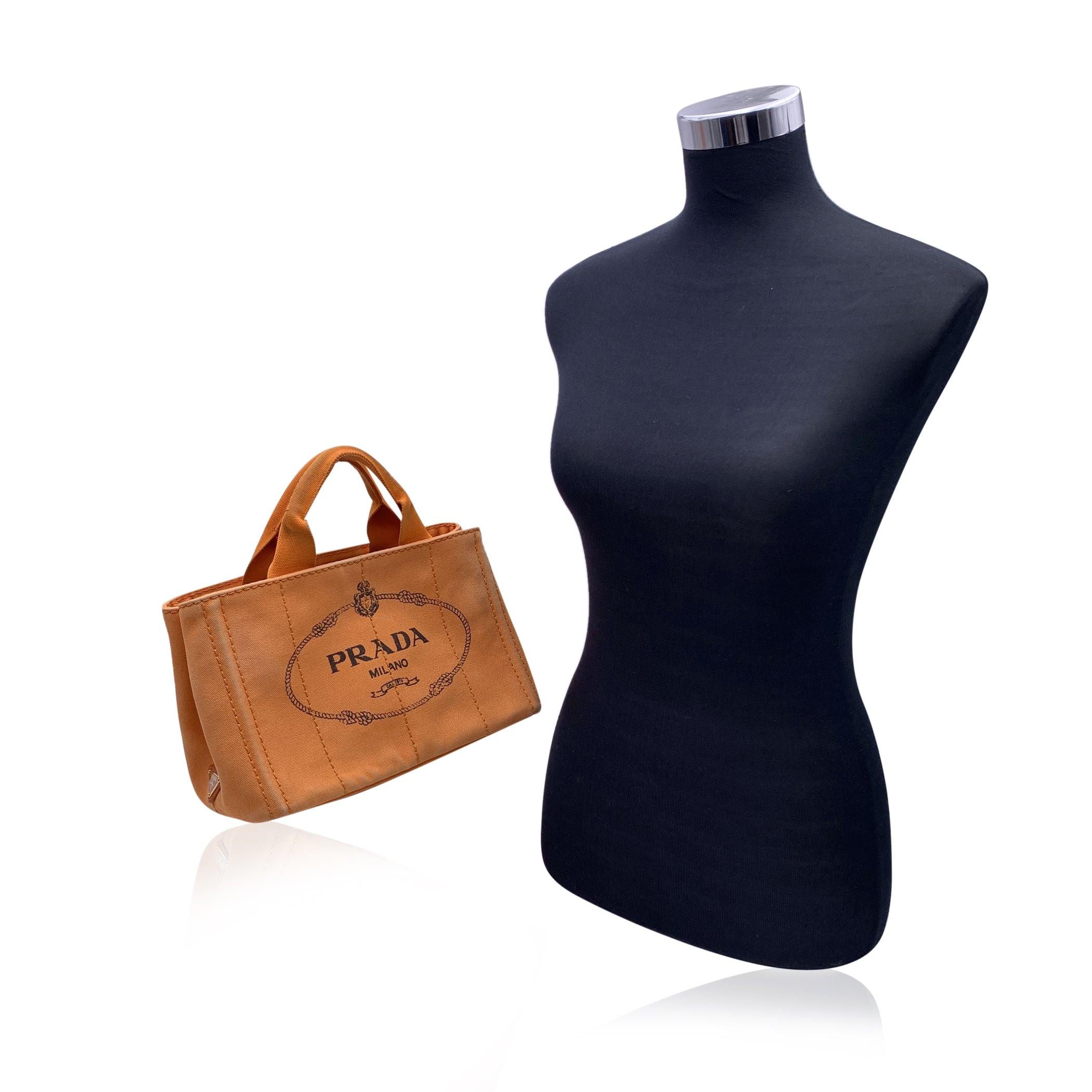 Prada Canapa small tote bag in orange hemp canvas with printed Prada logo and Prada triangle logo on the side. Canvas handles. Open top with side snap buttons on the sides. Canvas interior with 1 zip pocket and 3 open pockets and a light hemp