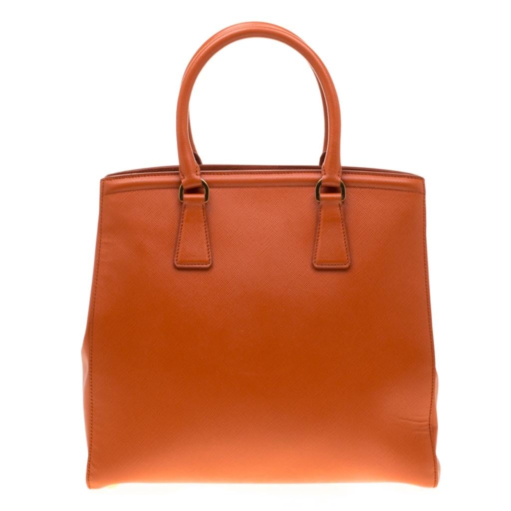 This sophisticated Parabole tote from Prada is crafted from Saffiano leather. The tote has an orange exterior featuring dual handles, a detachable shoulder strap and protective metal feet at the bottom. The fabric-lined interior houses an open