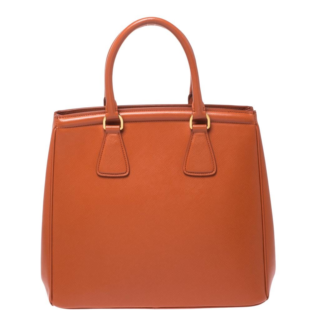 Masterfully created, this Prada tote bag is a style icon. Designed in a Saffiano Lux leather body in Italy, it exudes style and class in equal measures. This delightful orange piece is held by two top handles, is equipped with a brand logo,