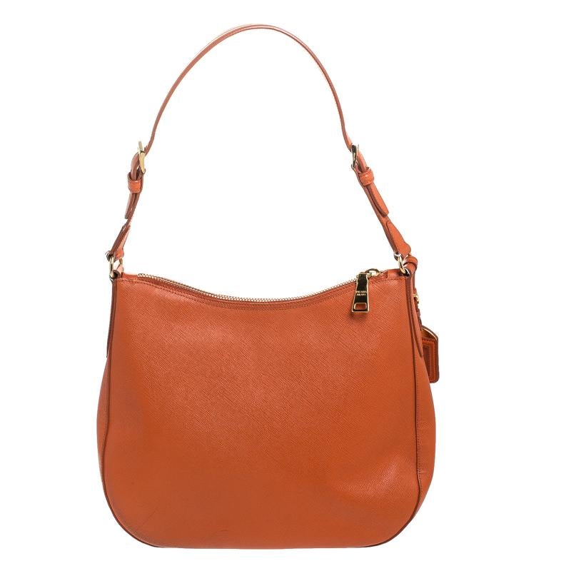 This stunning hobo bag comes from the house of Prada. It has been crafted in Italy and is made of luxurious Saffiano leather. It comes in a striking shade of orange and makes a statement. It is held by a single shoulder strap, features a top-zip