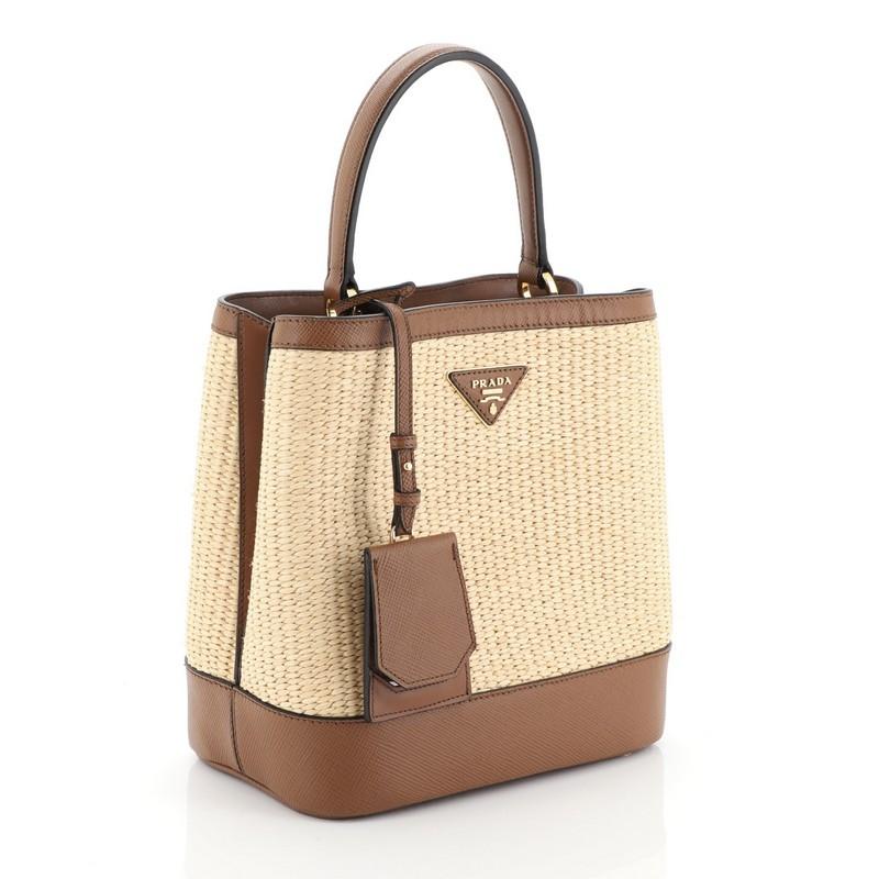 This Prada Panier Bucket Bag Straw and Leather Medium, crafted in brown and neutral leather, features a leather top handle and gold-tone hardware. Its wide open top showcases a brown leather interior divided into two compartments. 

Estimated Retail