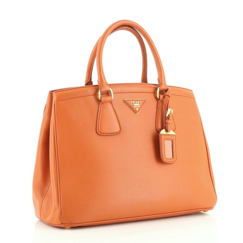 This Prada Parabole Handbag Saffiano Leather Medium, crafted in orange leather, features in dual rolled leather handles, protective base studs and gold-tone hardware. It opens to an orange fabric interior with middle zip compartment including a side