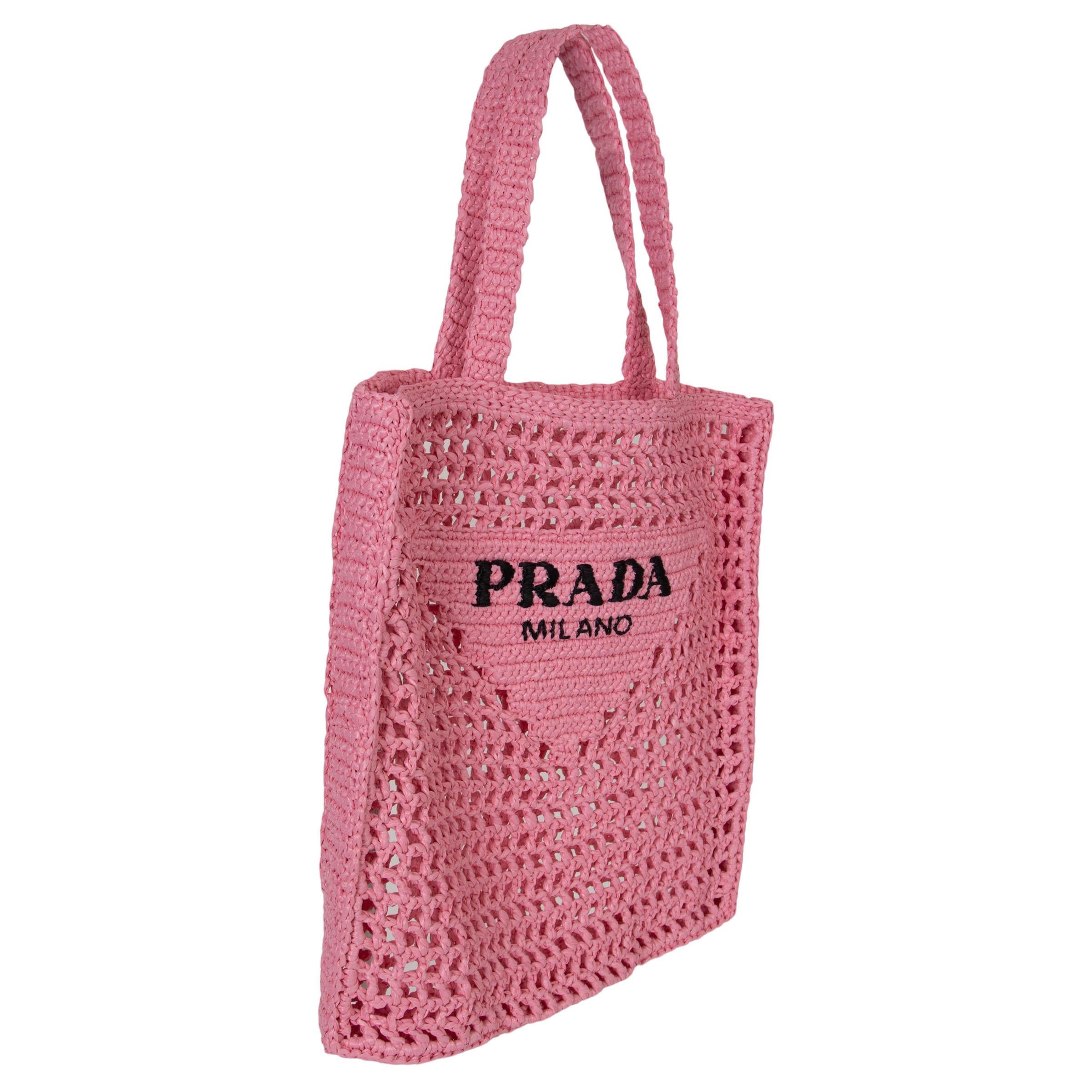 Este bolso tote 100% auténtico de Prada en hilo rosa pétalo con efecto rafia es una versión fresca y moderna de un diseño clásico. La parte delantera presenta una reinterpretación contemporánea del icónico motivo triangular de Prada, resaltado por