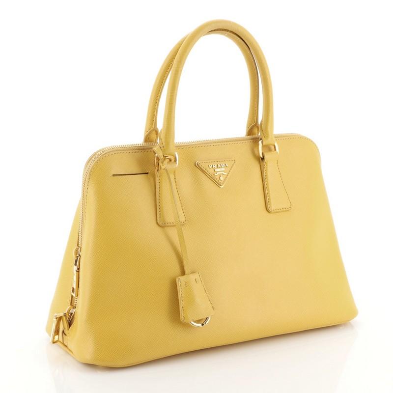 This Prada Promenade Bag Saffiano Leather Medium, crafted in yellow saffiano leather, features dual rolled handles, protective base studs, Prada logo at the center and gold-tone hardware. Its zip closure opens to a yellow fabric interior with side