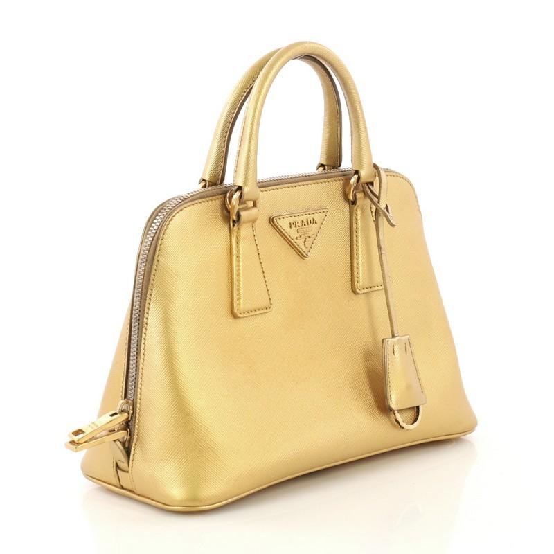 This Prada Promenade Bag Saffiano Leather Small, crafted from metallic gold saffiano leather, features dual rolled handles, triangle Prada logo, and gold-tone hardware. Its two-way zip closure opens to a gold fabric and leather interior with a
