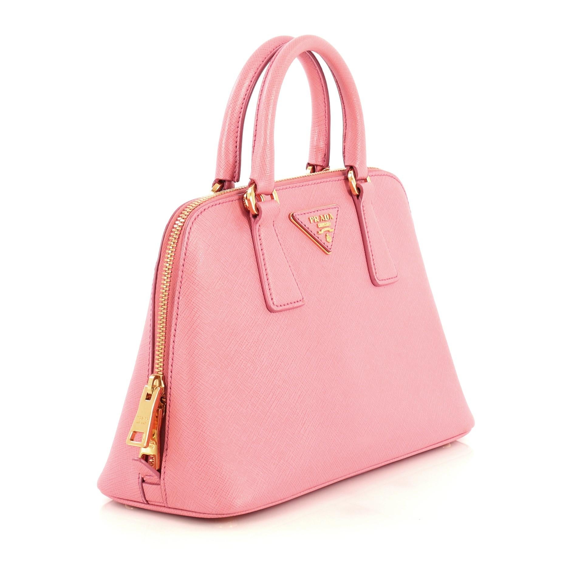 This Prada Promenade Bag Saffiano Leather Small, crafted from pink saffiano leather, features dual rolled handles, triangle Prada logo, and gold-tone hardware. Its two-way zip closure opens to a pink fabric interior with a center zip compartment and
