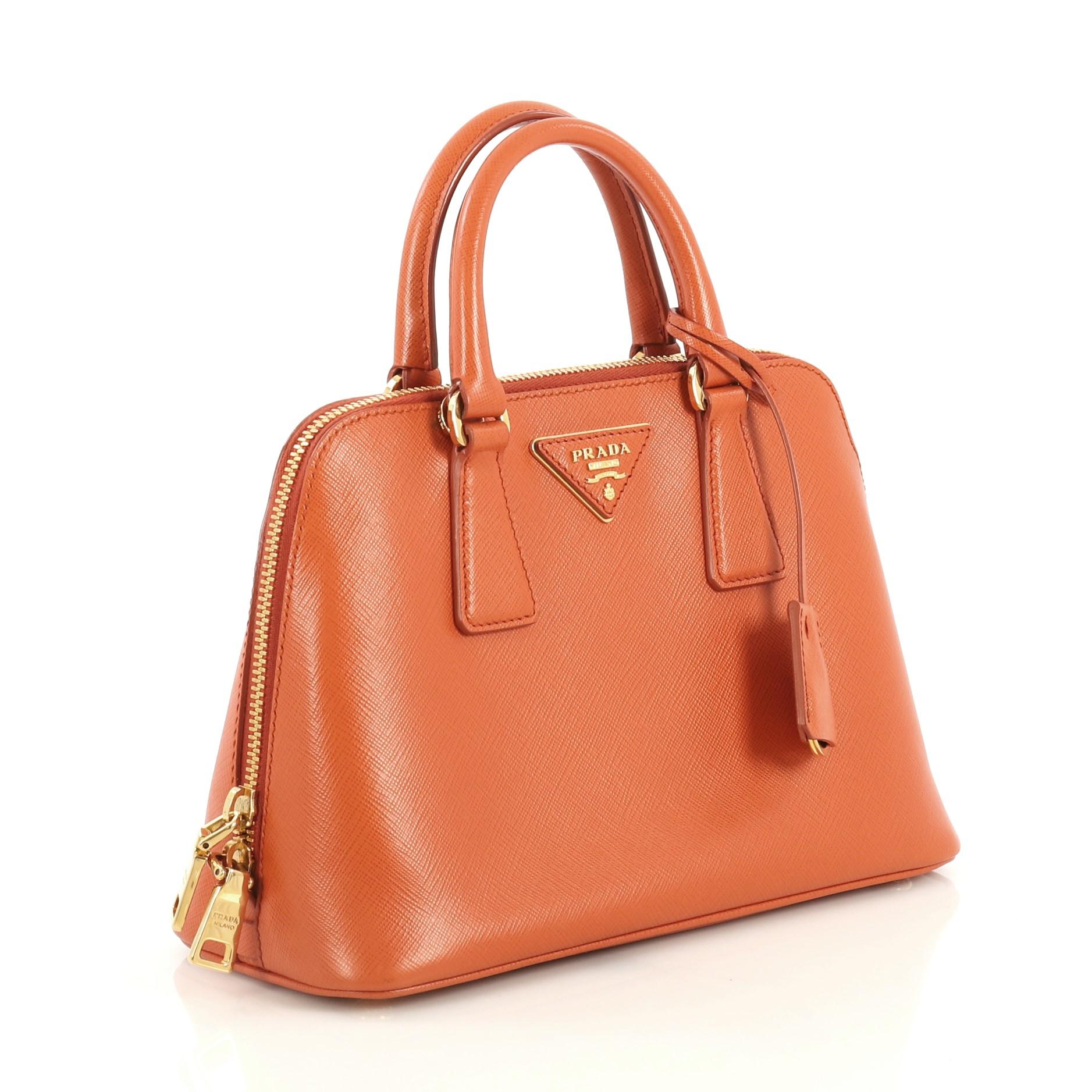 This Prada Promenade Bag Saffiano Leather Small, crafted from orange saffiano leather, features dual rolled handles, triangle Prada logo, and gold-tone hardware. Its two-way zip closure opens to an orange fabric interior with a center zip