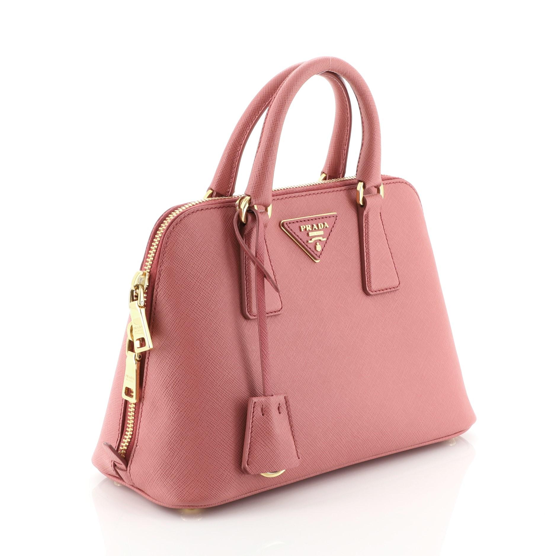 This Prada Promenade Bag Saffiano Leather Small, crafted from pink saffiano leather, features dual rolled handles, triangle Prada logo, and gold-tone hardware. Its two-way zip closure opens to a pink fabric interior with a center zip compartment and