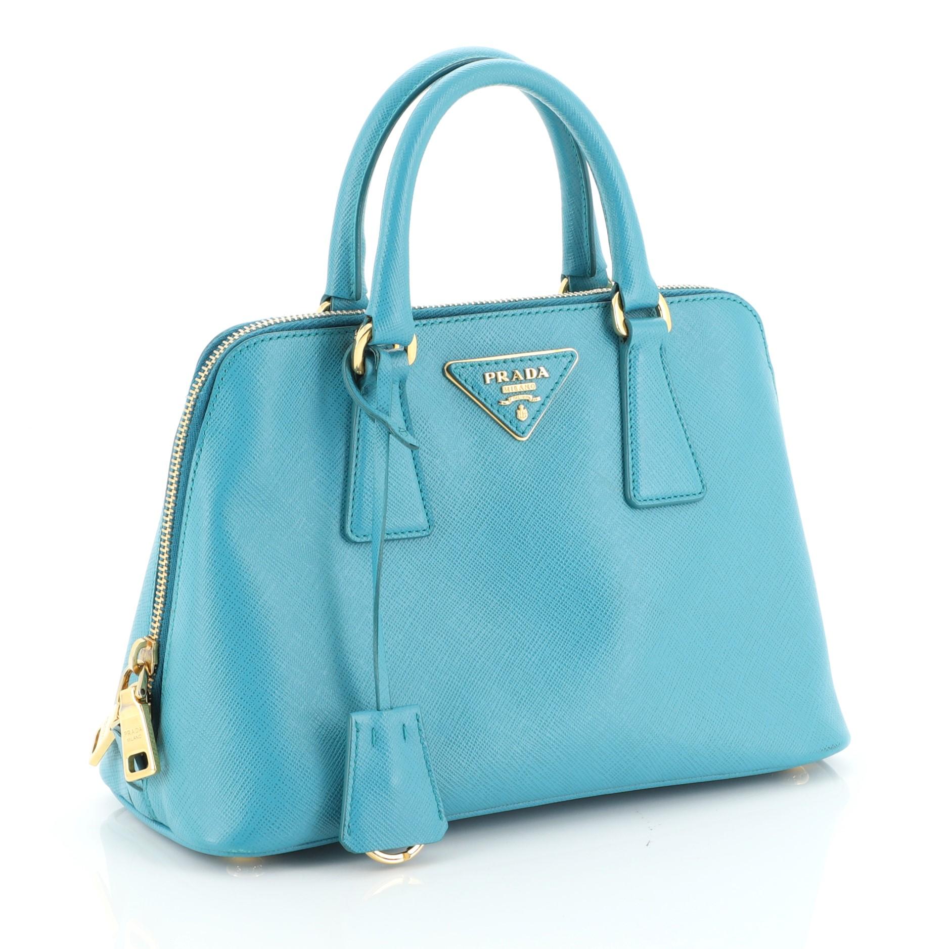 This Prada Promenade Bag Saffiano Leather Small, crafted from blue saffiano leather, features dual rolled handles, triangle Prada logo, and gold-tone hardware. Its two-way zip closure opens to a blue fabric interior with a center zip compartment and