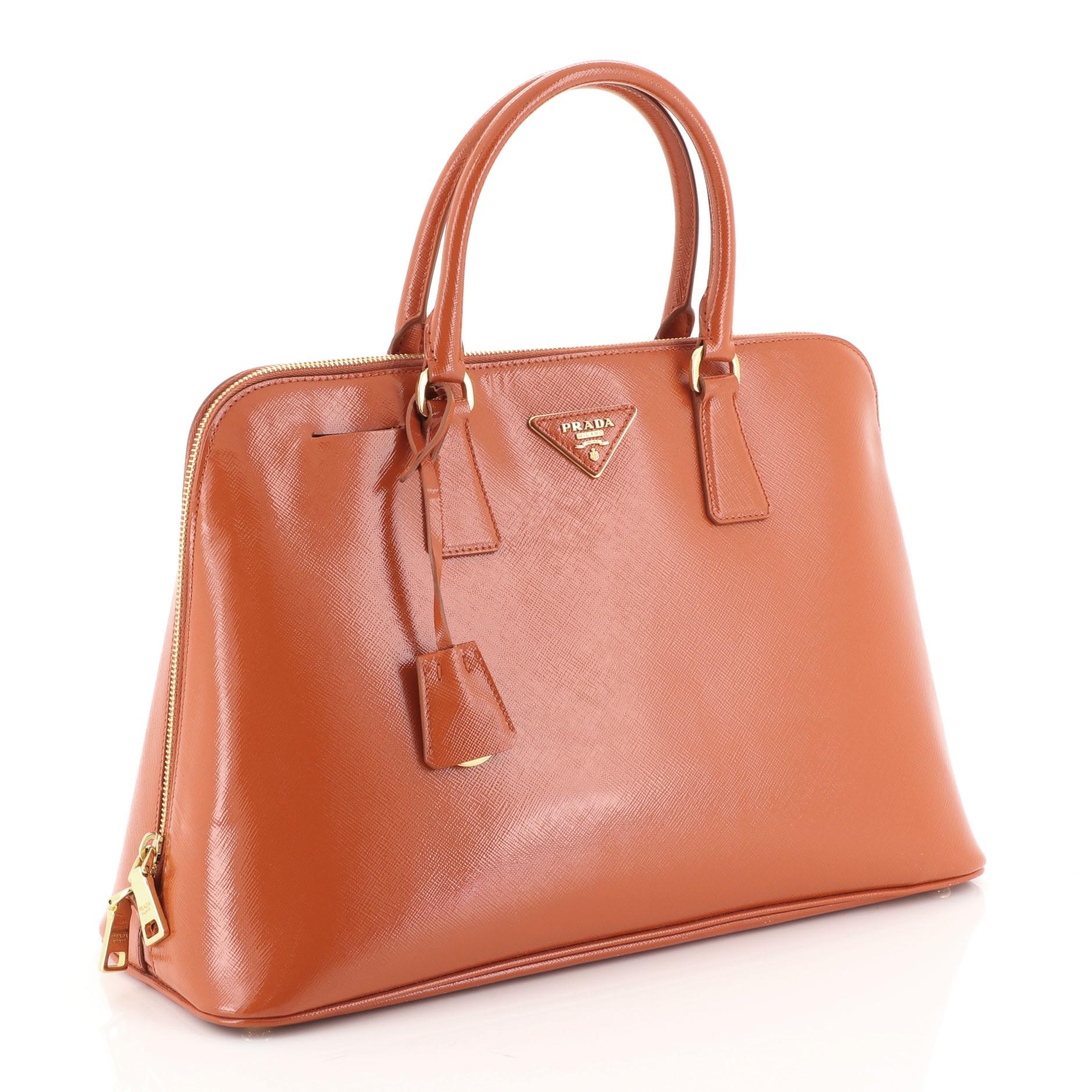 This Prada Promenade Bag Vernice Saffiano Leather Large, crafted from orange vernice saffiano leather, features dual rolled handles, triangle Prada logo, and gold-tone hardware. Its two-way zip closure opens to an orange fabric interior with a