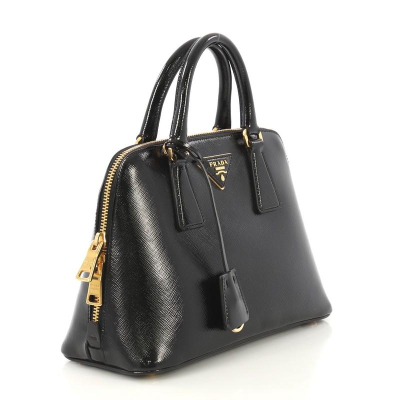 This Prada Promenade Bag Vernice Saffiano Leather Small, crafted from black vernice saffiano leather, features dual rolled handles, triangle Prada logo, and gold-tone hardware. Its two-way zip closure opens to a black fabric and leather interior