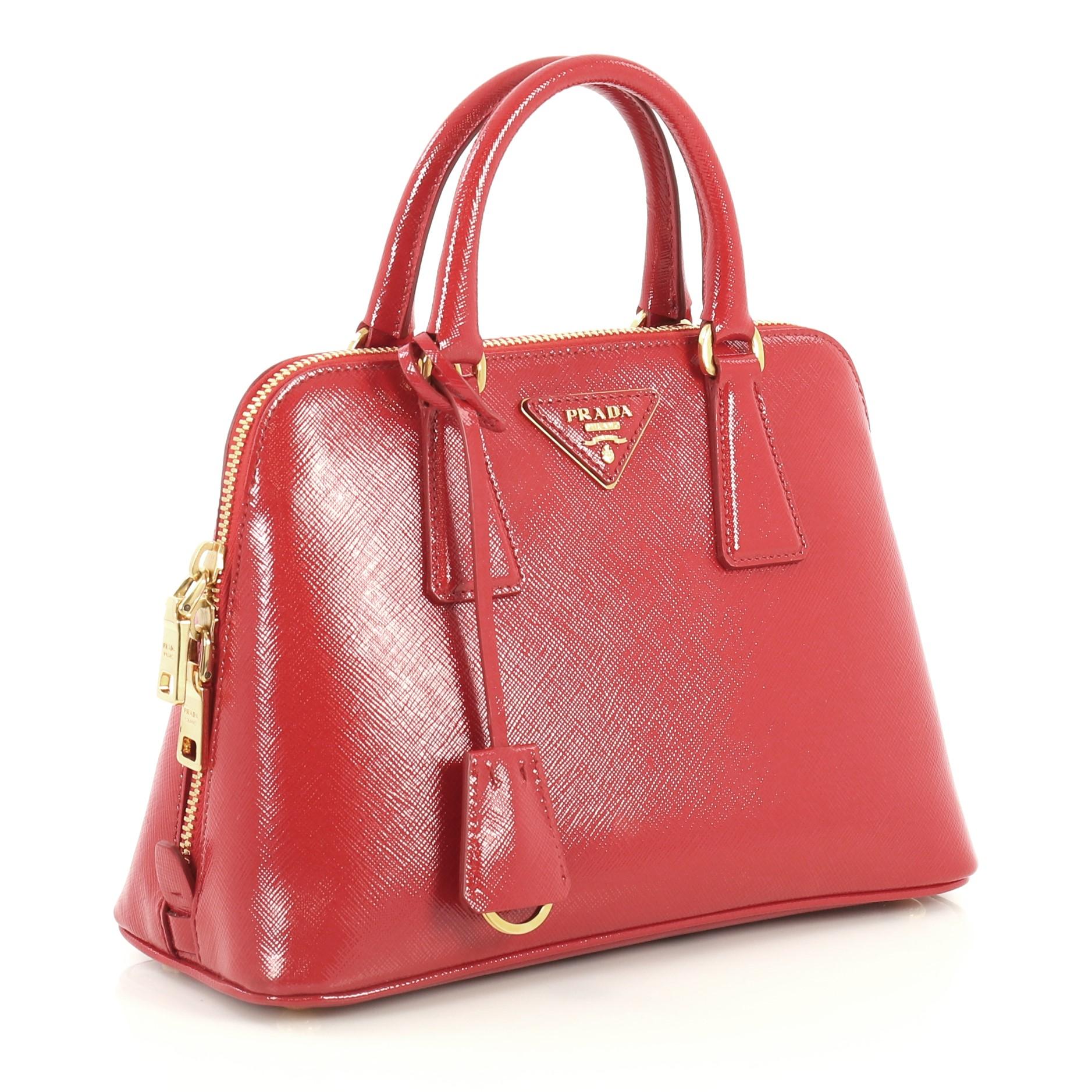 This Prada Promenade Bag Vernice Saffiano Leather Small, crafted from red vernice saffiano leather, features dual rolled handles, protective base studs and gold-tone hardware. Its zip closure opens to a red fabric interior divided into two