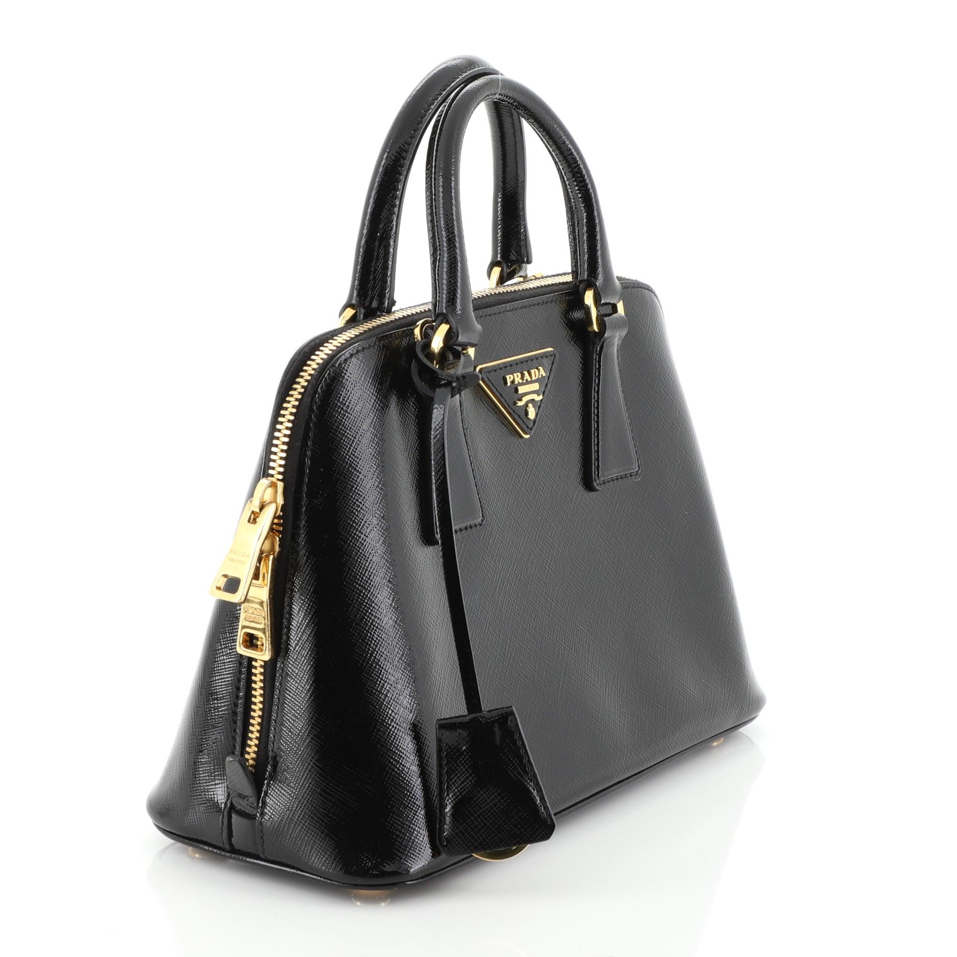 This Prada Promenade Bag Vernice Saffiano Leather Small, crafted from black vernice saffiano leather, features dual rolled handles, protective base studs and gold-tone hardware. Its zip closure opens to a black fabric interior divided into two