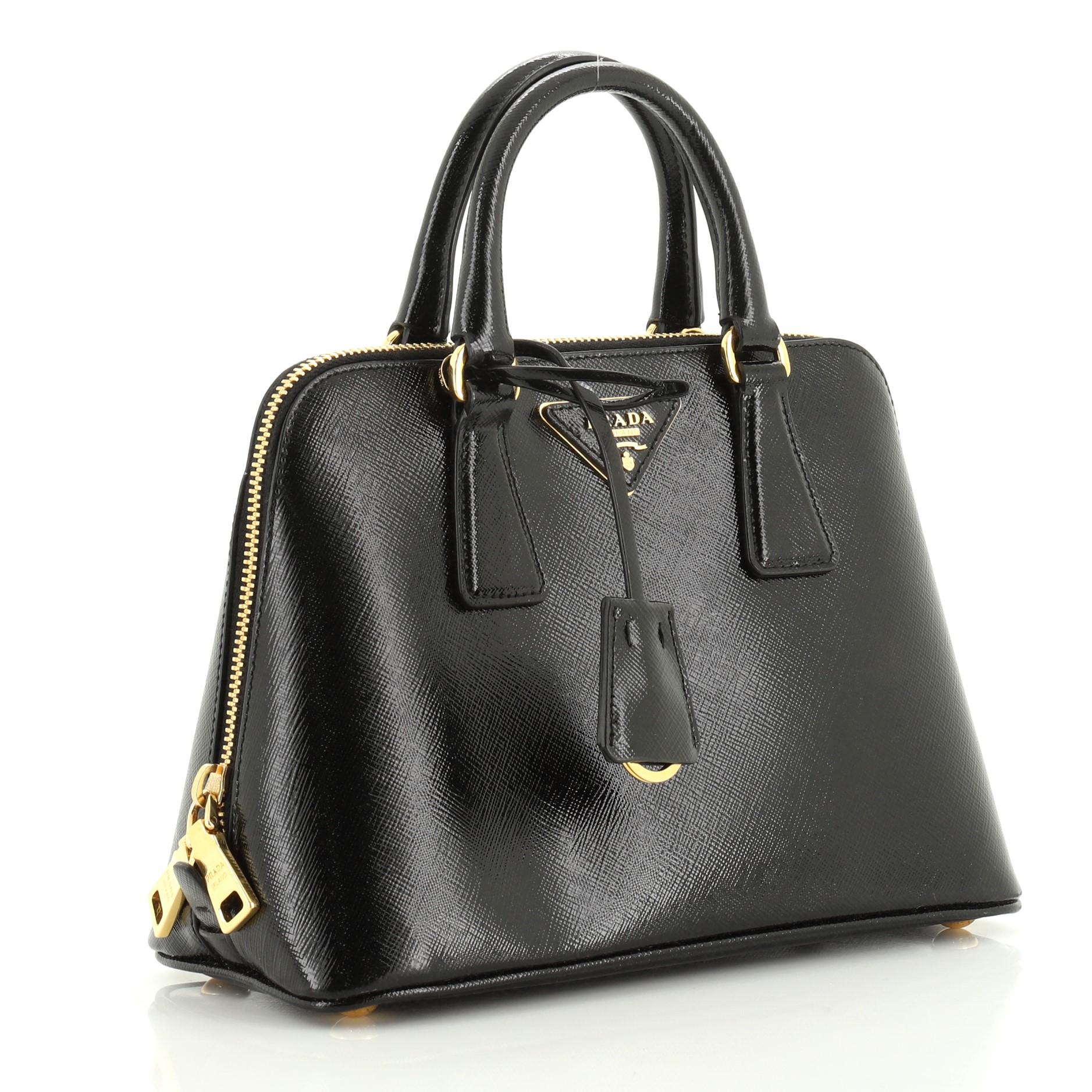 This Prada Promenade Bag Vernice Saffiano Leather Small, crafted from black vernice saffiano leather, features dual rolled handles, triangle Prada logo, and gold-tone hardware. Its two-way zip closure opens to a black fabric interior with a center