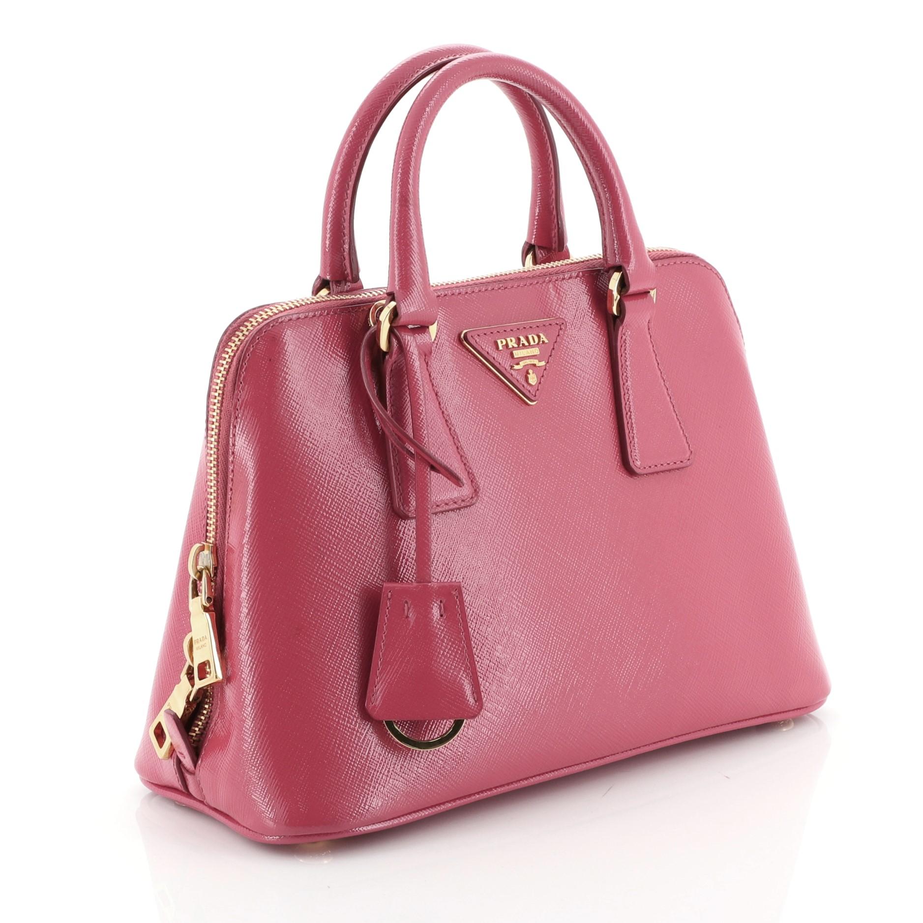 This Prada Promenade Handbag Vernice Saffiano Leather Small, crafted from pink vernice saffiano leather, features dual rolled handles, triangle Prada logo, and gold-tone hardware. Its two-way zip closure opens to a pink fabric interior with a center