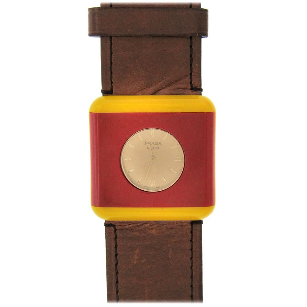 Prada definiert den Stil von Uhren mit ihrem kühnen, quadratischen Gehäuse in Rot und Gelb auf einem extrabreiten Lederband mit Schnalle.  Die Quarzuhr misst 45 mm im Quadrat, hat verdeckte Bandanstöße, ein schweres braunes Lederband und ein