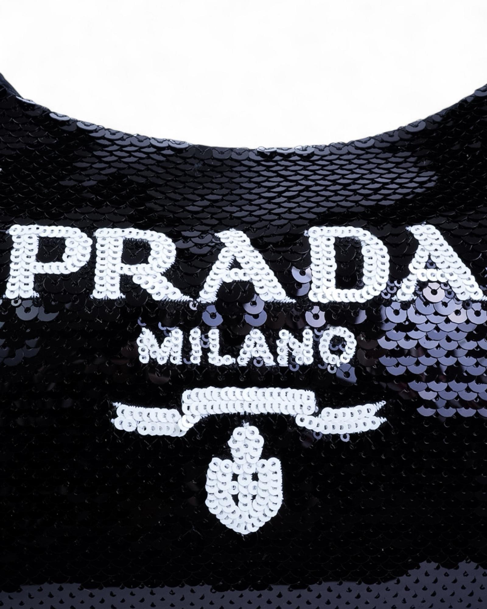 Sac signé Prada, modèle Re-Edition 2000, en paillettes noires avec détails argentés. Le sac est doté d'une fermeture à glissière sur le dessus, tandis que l'intérieur est doublé d'un tissu portant le logo, offrant ainsi suffisamment d'espace pour
