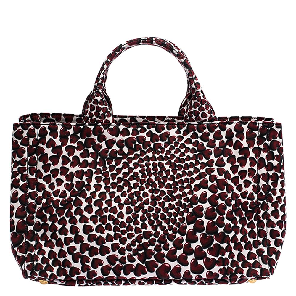One of the most iconic designs from the house of Prada, this Canapa Tote bag is great to wear through the day or at your vacations whilst never compromising on style. Covered in a heart print, this red-hued bag features two handles, a spacious
