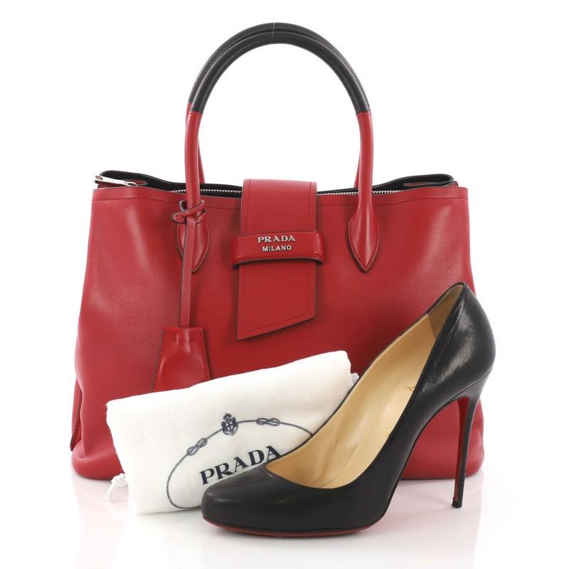 This Prada Ribbon Tote City Calfskin Medium, crafted in red calfskin leather, features dual two tone leather handles, protective base studs, and silver-tone hardware. Its belt tab closure opens to a black leather interior with two open compartments
