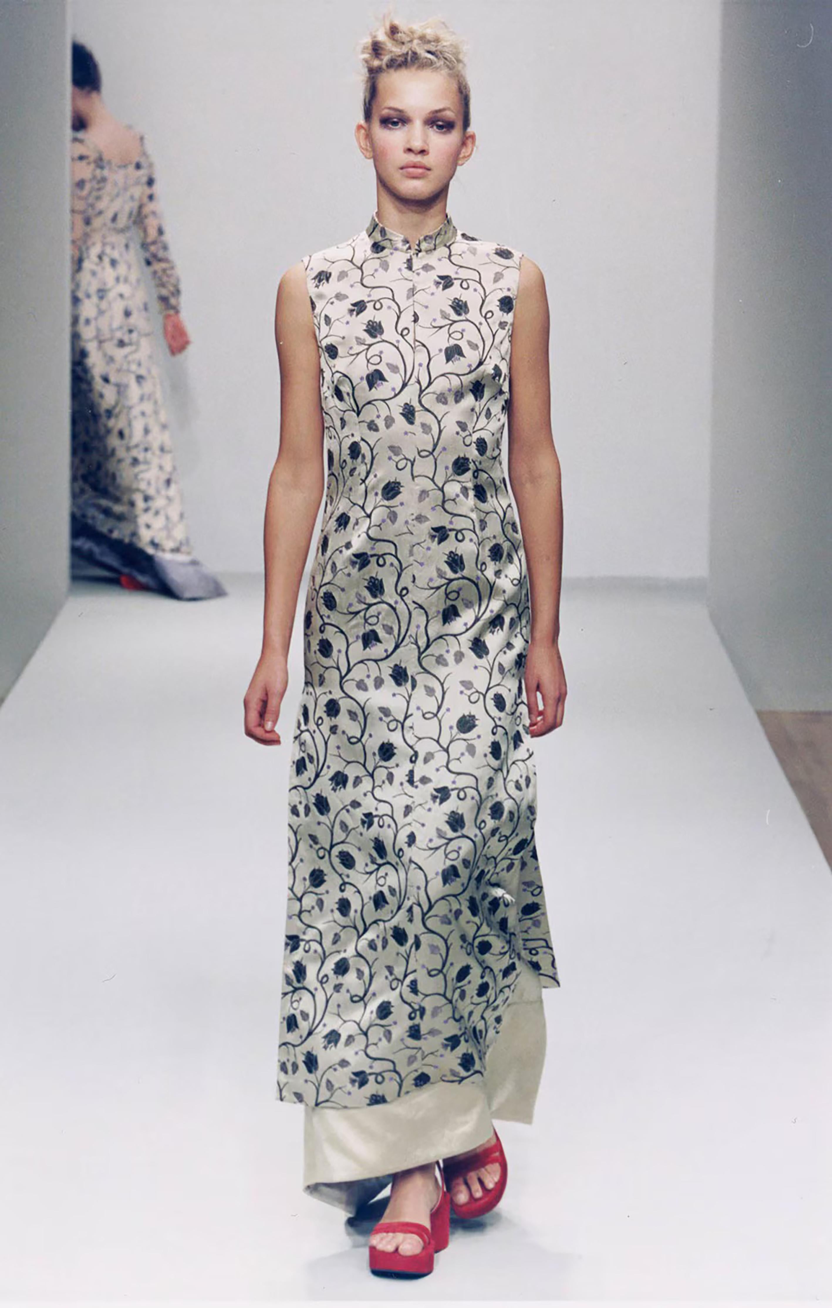 Il mini abito cheongsam in seta floreale Eleganza di Prada, realizzato in seta delicata, vanta un raffinato colletto alla mandarina che sottolinea la scollatura con una sottile eleganza. Adornato da un'intricata stampa floreale, il pezzo emana un