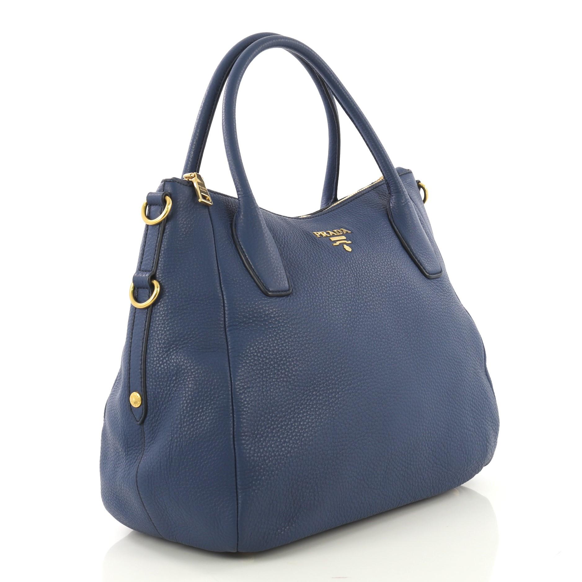 This Prada Sacca 2 Manici Convertible Tote Vitello Daino Medium, crafted from blue vitello daino leather, features dual rolled top handles, raised Prada Milano logo at its center, protective base studs, and gold-tone hardware. Its zip closure opens