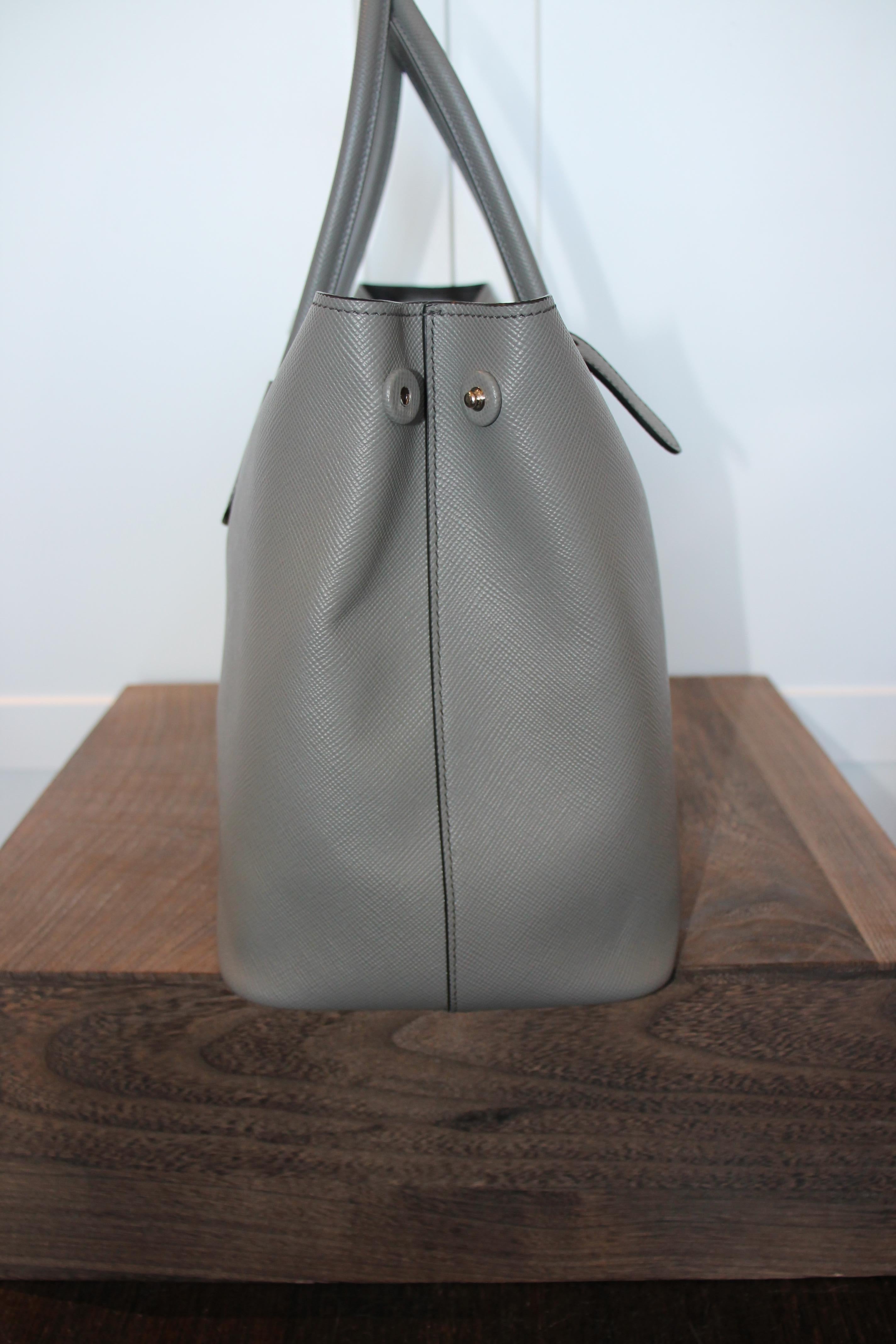 Gray grained leather. Silver-tone hardware. Opened top with fold over closure with buckle closures. Dual top rolled handles. Prada logo at front. Features detachable dog tag.