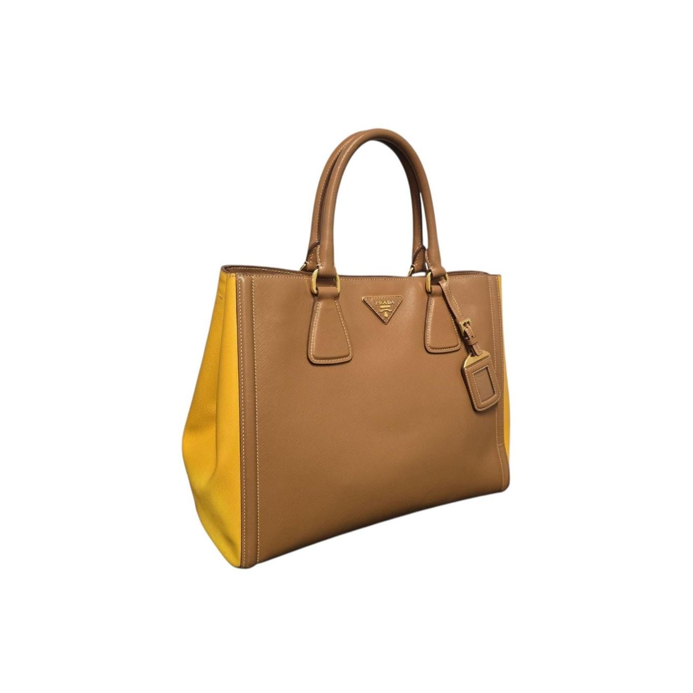 It features a bicolor design, typically brown/caramel with yellow side panels, and is made from Prada's signature Saffiano cross-grain leather, rolled leather top handles, an optional and adjustable shoulder strap, and a matching luggage tag. It is