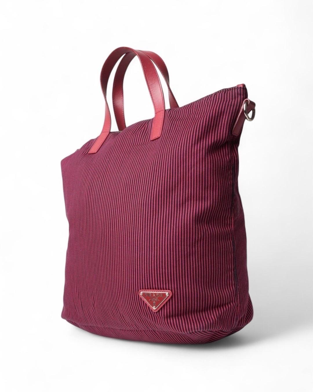 Borsa firmata Prada, modello Shopper Vertical nella misura medium, realizzata in tessuto con fantasia a righe e rifiniture in pelle rossa e hardware argento. Dotada de un cierre superior de cremallera, internamente revestida de tejido logado tono