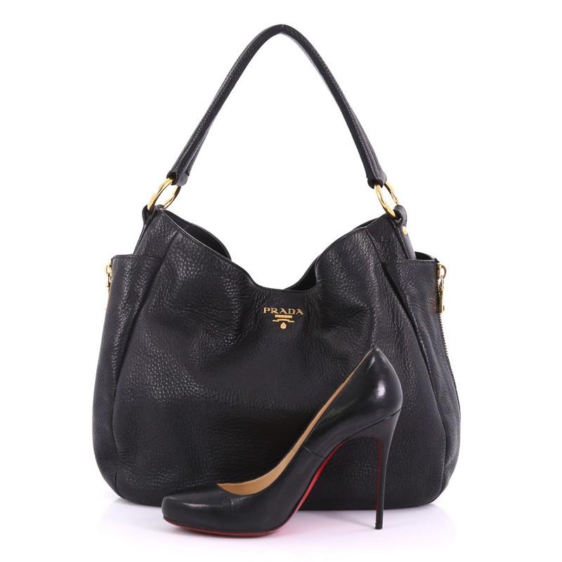 This Prada Side Pocket Zip Hobo Vitello Daino Large, crafted from black vitello daino leather, features a flat leather handle, two exterior side flat pockets with side zipper details, Prada Milano logo, and gold-tone hardware. Its snap button