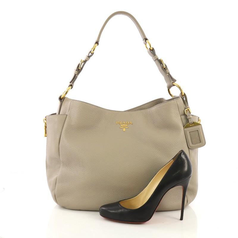 This Prada Side Pocket Zip Hobo Vitello Daino Large, crafted from gray vitello daino leather, features a flat leather handle, two exterior side flat pockets with side zipper details, Prada Milano logo, and gold-tone hardware. Its snap button closure