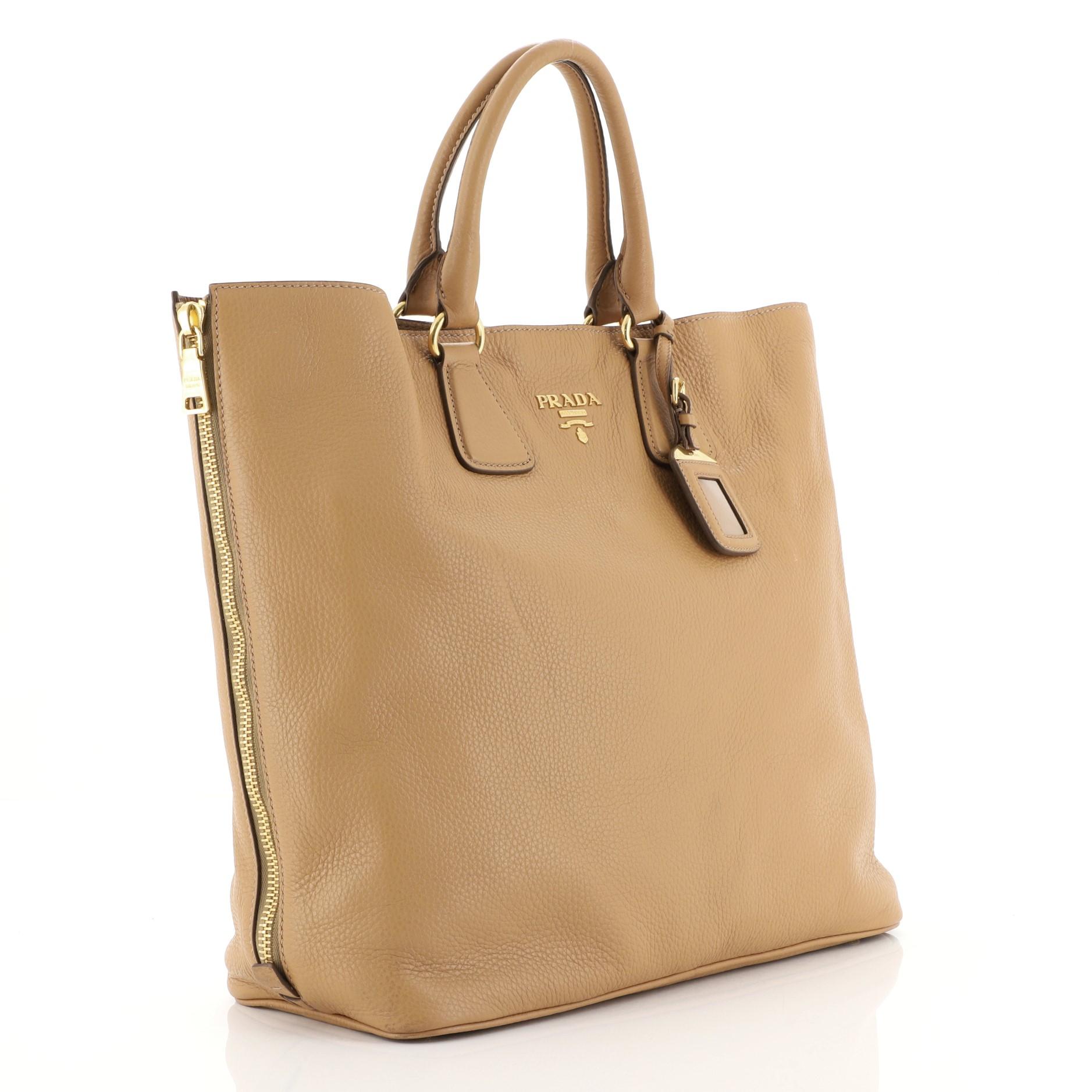 This Prada Side Zip Convertible Shopper Tote Vitello Daino Large, crafted from brown vitello daino leather, features dual rolled handles, raised Prada logo at its center, and gold-tone hardware. Its snap closure opens to a brown fabric interior with