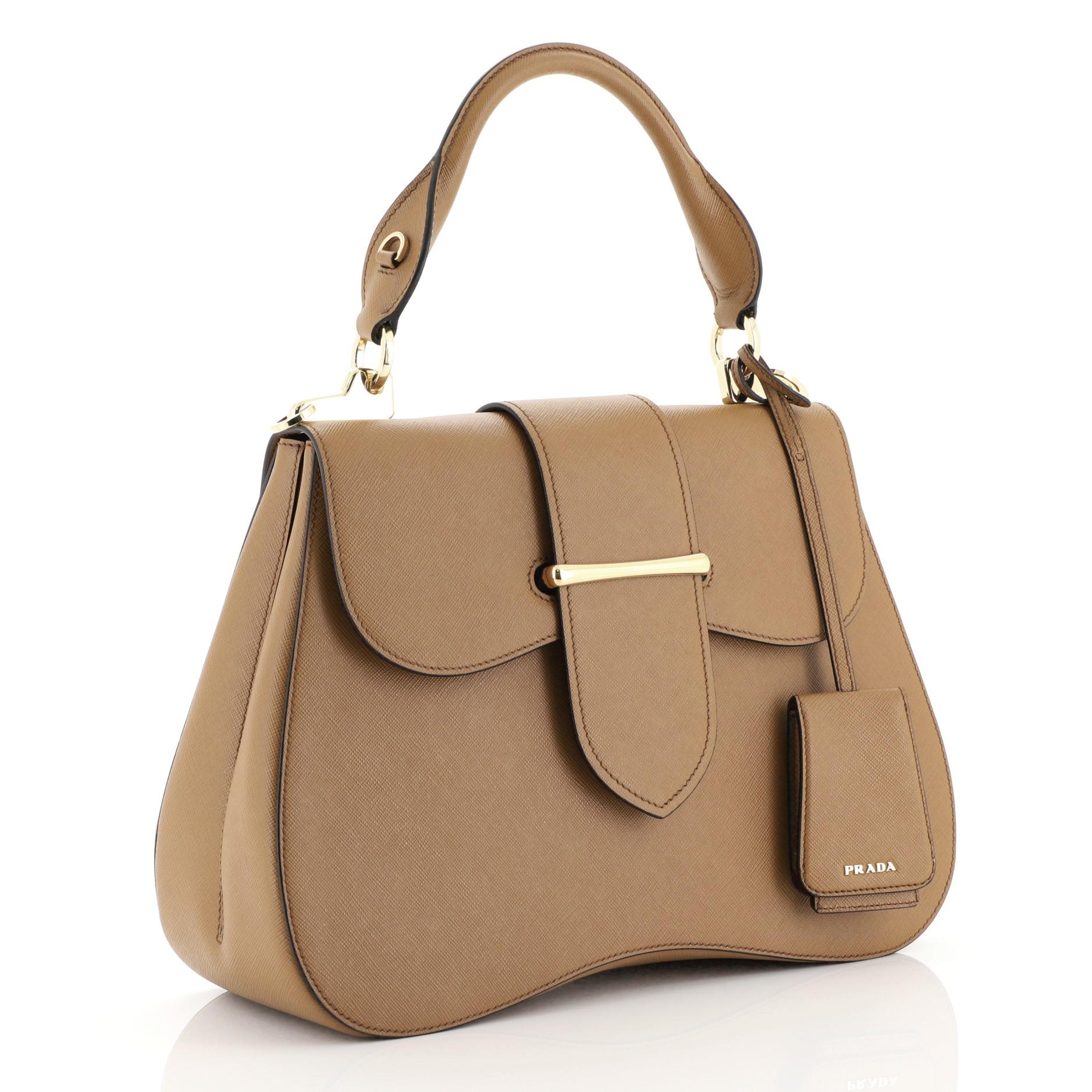 This Prada Sidonie Top Handle Bag Saffiano Leather Large, crafted from brown saffiano leather, features a rolled top handle and gold-tone hardware. Its flap opens to a red leather interior with side zip and slip pockets. 

Estimated Retail Price: