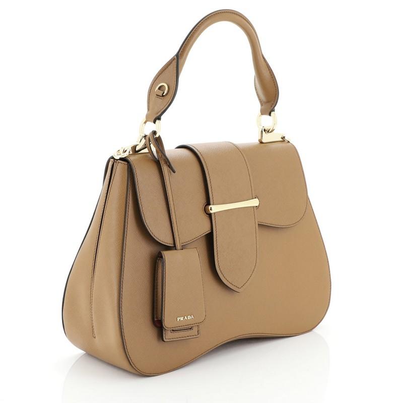 This Prada Sidonie Top Handle Bag Saffiano Leather Large, crafted from brown saffiano leather, features a rolled top handle and gold-tone hardware. Its flap opens to a red leather interior with side zip and slip pockets. 

Condition: Excellent.